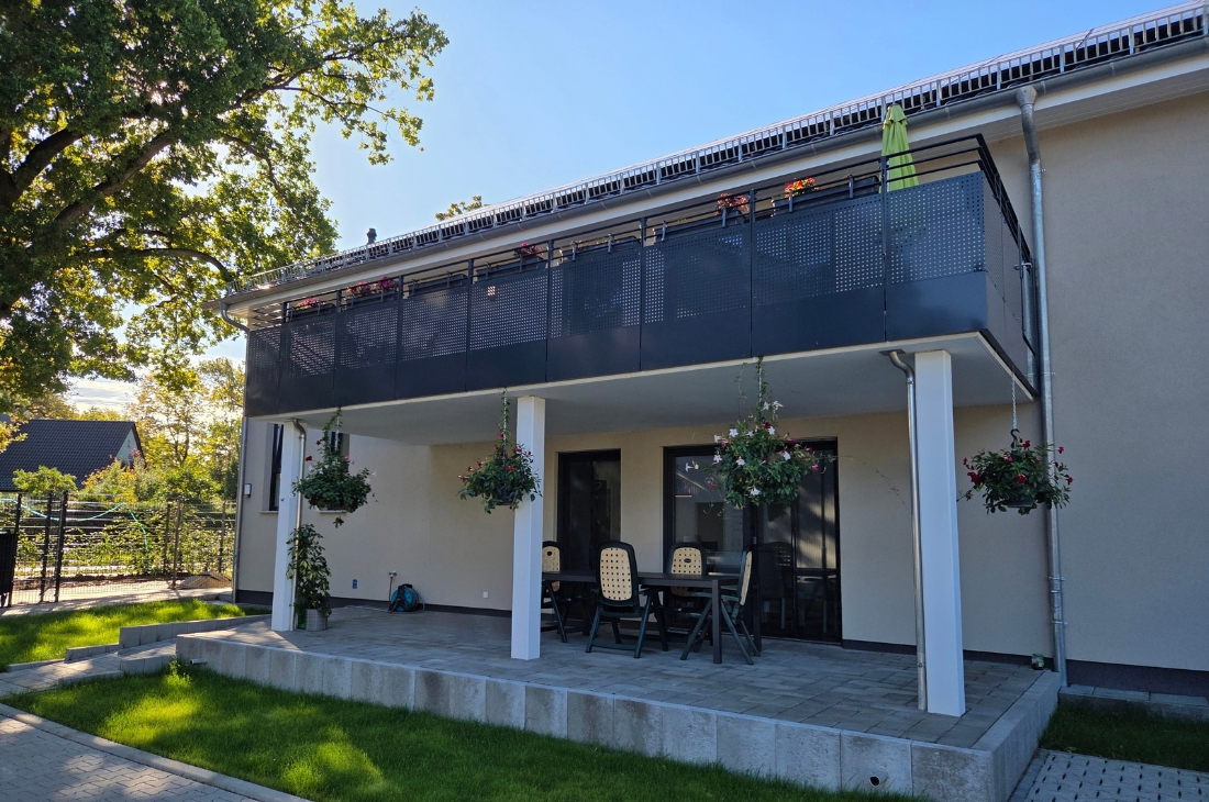 Schräge Ansicht der Terrasse mit Sitzgruppe unter dem Balkon, getragen von weißen Stützen mit hängenden Blumenampeln. Links ein großer Laubbaum und ein Zaun, rechts ein Fallrohr an der beigen Fassade. Die Terrasse ist durch eine Betonblockkante vom Rasen abgesetzt.
