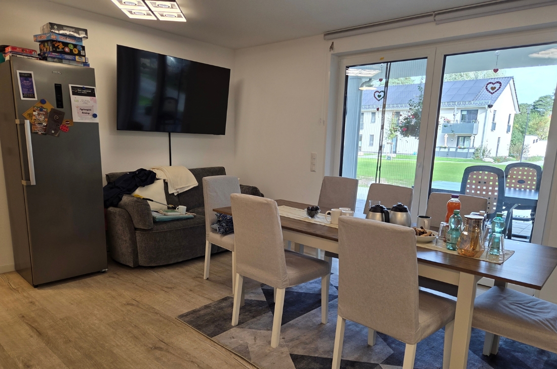 Wohn- und Essbereich mit Esstisch aus Holz, hellgrau gepolsterten Stühlen und gedecktem Tisch mit Kannen, Tassen und Getränken. Im Hintergrund ein Sofa, ein an der Wand montierter Fernseher und ein Edelstahl-Kühlschrank, auf dem Gesellschaftsspiele gestapelt sind. Durch die bodentiefen Fenster sieht man Rasen und ein Nachbargebäude.
