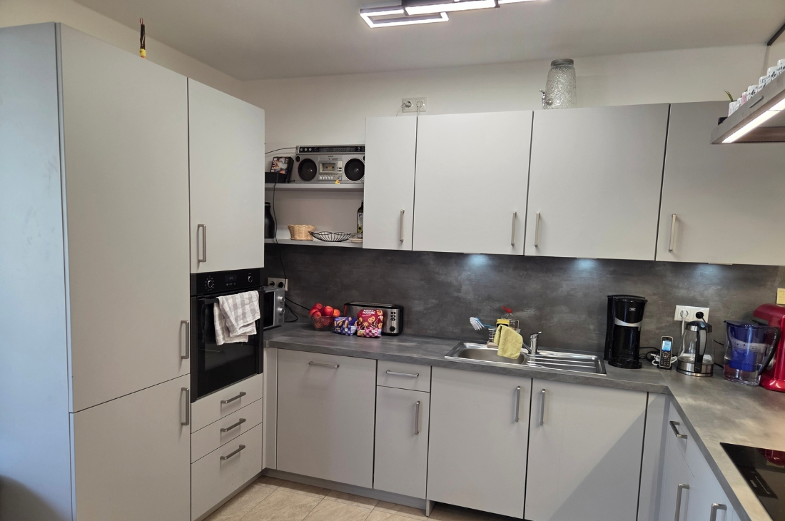 Moderne L-förmige Küche mit hellgrauen Fronten, dunkler Arbeitsplatte in Steinoptik und Spüle. Auf der Arbeitsplatte stehen Kaffeemaschine, Wasserkocher, Toaster und eine Obstschale, im offenen Regal ein Radiorekorder. Im Vordergrund links der Backofen im Hochschrank, rechts das Kochfeld.