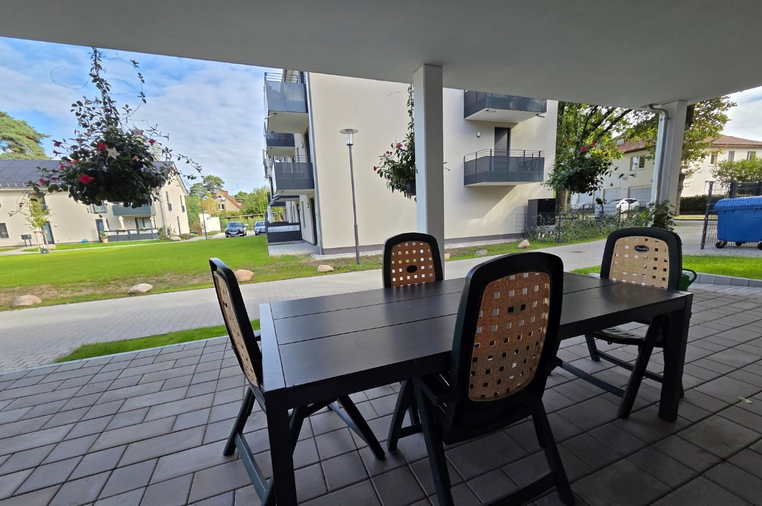 Blick von der überdachten Terrasse auf die Wohnanlage. Im Vordergrund ein dunkler Tisch mit Stühlen, dahinter Rasen, ein Weg und weitere Wohngebäude mit Balkonen. Rechts stehen ein blauer Müllcontainer und ein Zaun, links hängt eine blühende Blumenampel.