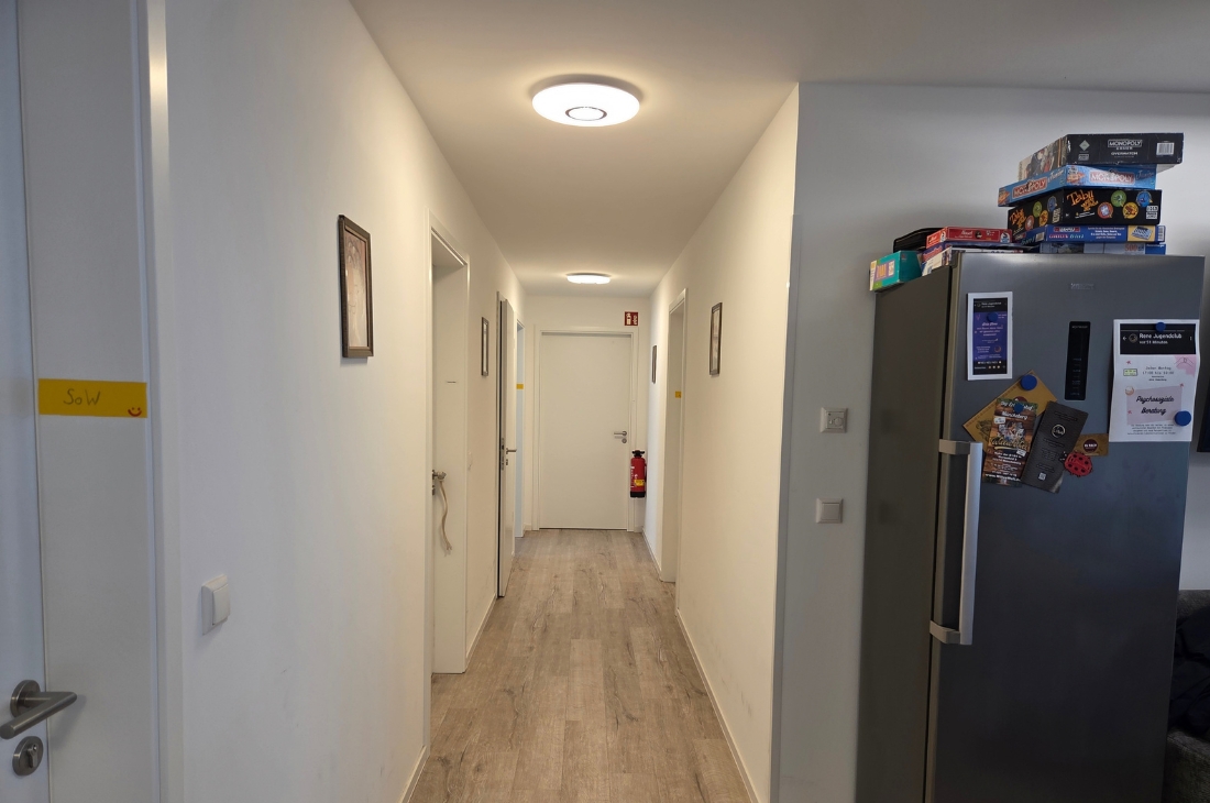 Heller Flur mit Holzoptik-Boden und mehreren weißen Türen, an den Wänden hängen kleine gerahmte Bilder. Am Ende des Flurs ist ein Feuerlöscher zu sehen, rechts ein Edelstahl-Kühlschrank mit Aushängen und gestapelten Gesellschaftsspielen. 