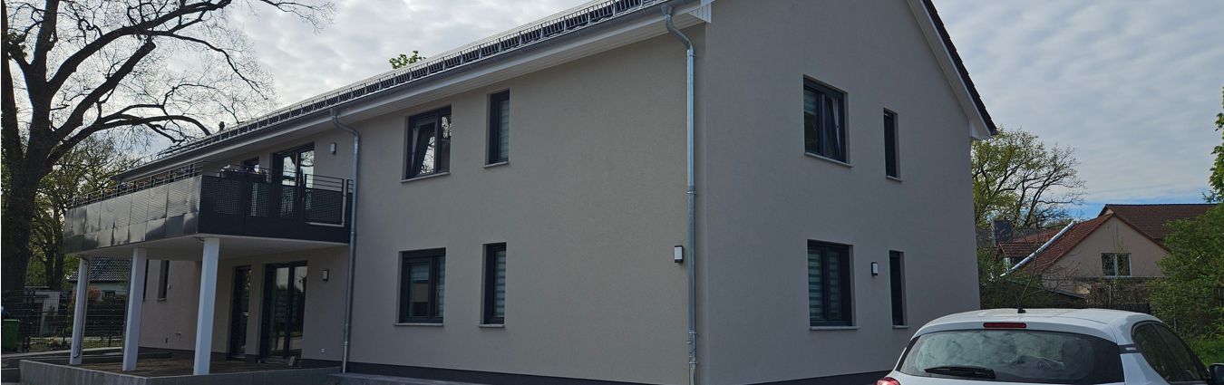 Modernes, zweigeschossiges Wohnhaus mit heller Putzfassade und dunklen Fensterrahmen. Auf der linken Seite befindet sich ein überdachter Balkon mit Sichtschutz; darunter liegt eine Terrasse mit Stützen. Rechts ist ein Teil eines geparkten Autos zu sehen. Im Hintergrund stehen weitere Einfamilienhäuser sowie Bäume unter einem leicht bewölkten Himmel.