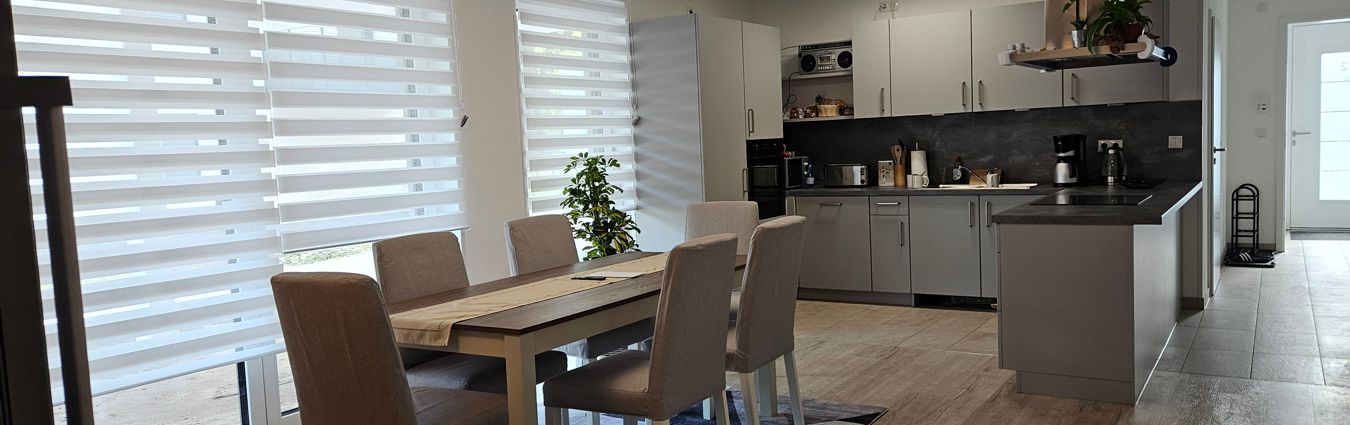 Heller, offener Wohn- und Essbereich mit moderner Küche. Im Vordergrund steht ein Esstisch aus Holz mit vier gepolsterten Stühlen; dahinter befinden sich große Fenster mit halbtransparenten Doppelrollos. Im Hintergrund ist eine L-förmige Einbauküche mit hellen Schränken, dunkler Arbeitsplatte, Kaffeemaschine und weiteren Küchengeräten zu sehen. Rechts führt ein Flur zu einer Tür, und eine Kücheninsel trennt den Kochbereich optisch vom Essbereich.
