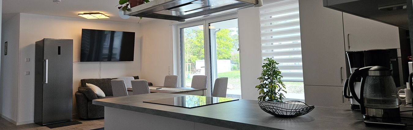 Offener Wohn- und Essbereich mit moderner Küche und direktem Zugang zur Terrasse. Im Vordergrund befindet sich eine Kücheninsel mit Kochfeld und dunkler Arbeitsplatte; rechts steht eine Kaffeemaschine. Dahinter steht ein Esstisch mit mehreren gepolsterten Stühlen vor großen Fenster- und Terrassentüren mit halbtransparenten Rollos. Im linken Hintergrund sind ein Sofa, ein großer Flachbildfernseher an der Wand und ein freistehender Kühlschrank zu sehen. Natürliches Licht fällt durch die Glasflächen und zeigt den grünen Außenbereich.