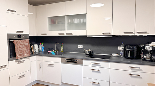 Moderne Küche mit weißen Hochglanzfronten, grauer Arbeitsplatte und verschiedenen Küchengeräten, darunter Backofen, Spülmaschine und Kaffeemaschine.