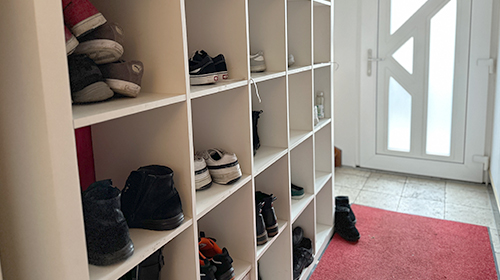 Flur mit einem weißen Regalsystem voller Schuhe, rotem Teppich und einer weißen Eingangstür mit Milchglas.