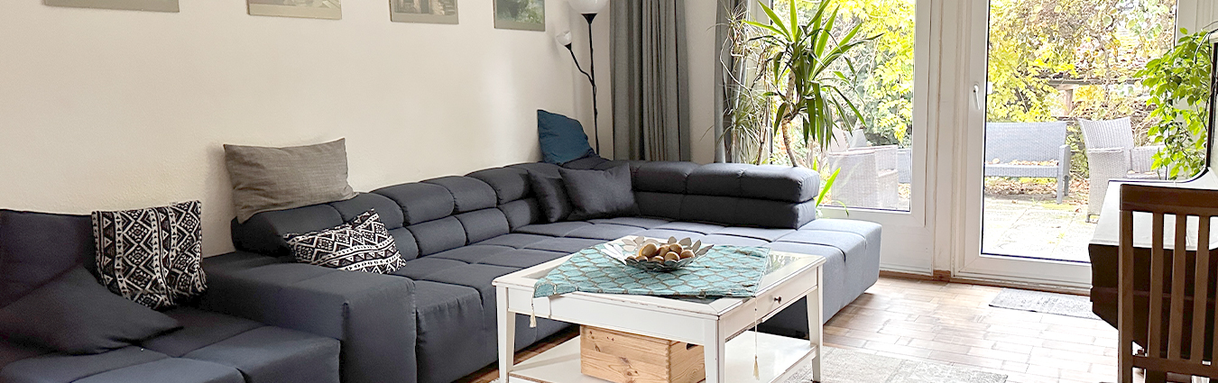 Helles Wohnzimmer mit großer grauer Eckcouch, weißem Couchtisch, Zimmerpflanzen und bodentiefen Fenstern mit Blick auf eine Terrasse und Gartenmöbel.