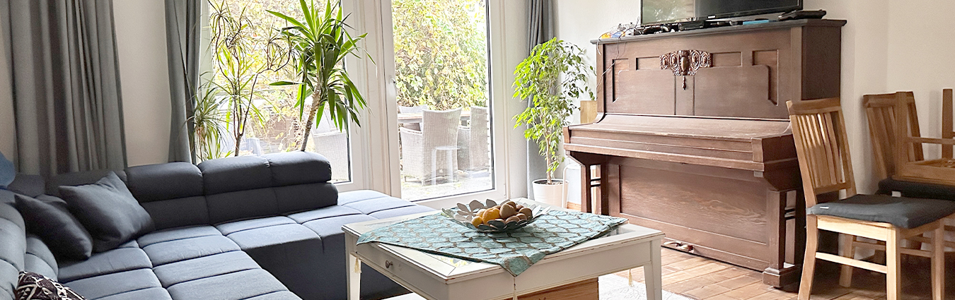 Helles Wohnzimmer mit großer grauer Eckcouch, weißem Couchtisch, mehreren Zimmerpflanzen und einem braunen Klavier mit Holzstühlen daneben, vor bodentiefen Fenstern mit Blick auf die Terrasse.
