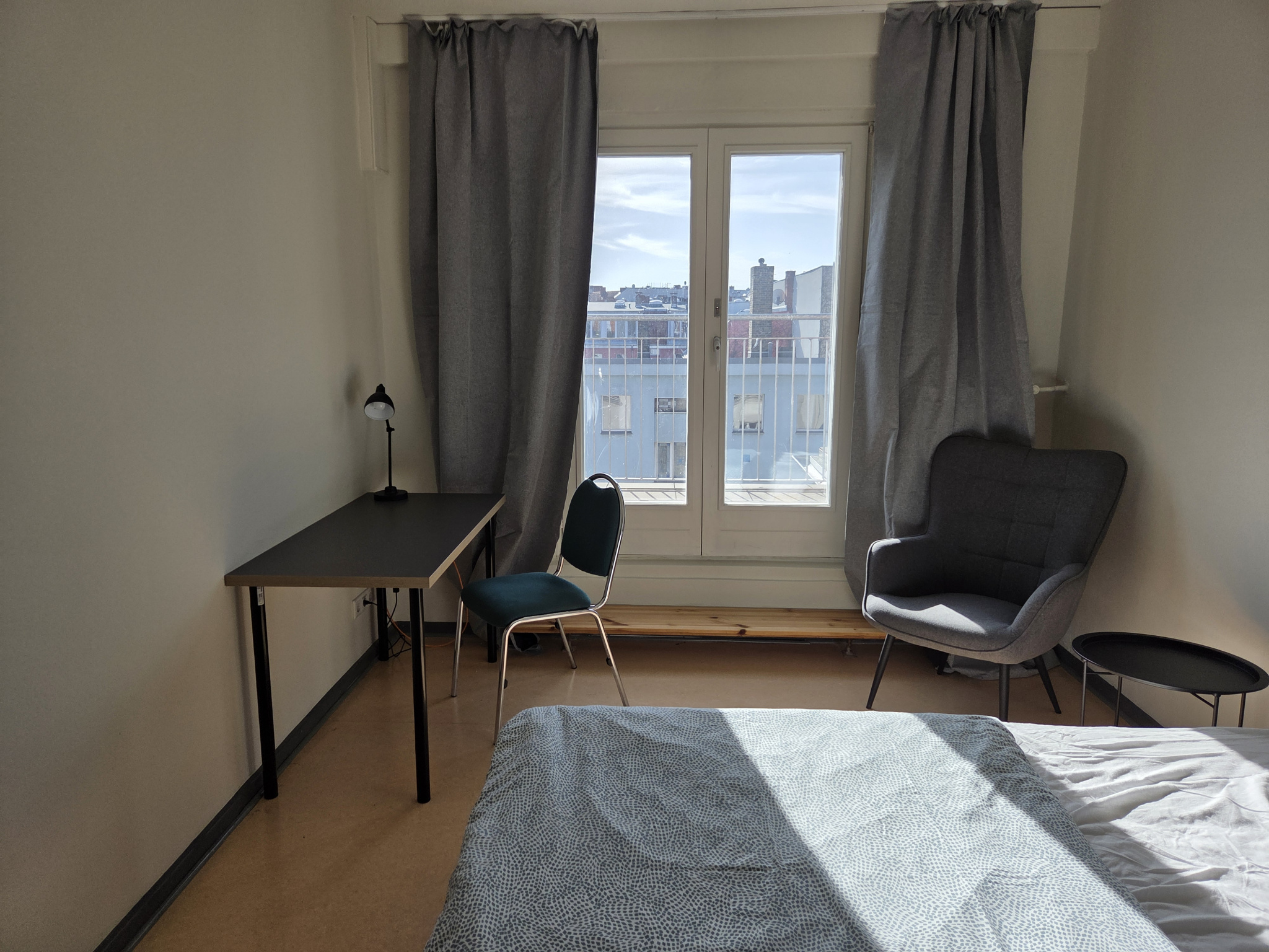 Ein helles Zimmer mit Terassentür, Bett, grauen Vorhängen, einem kleinen Sessel, Beistelltisch, einem Schreibtisch mit Stuhl und Lampe.