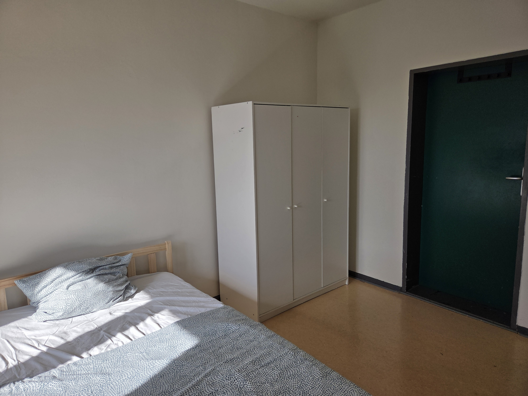 Ein helles Zimmer mit einem Bett, einer Eingangstür und einem Kleiderschrank.