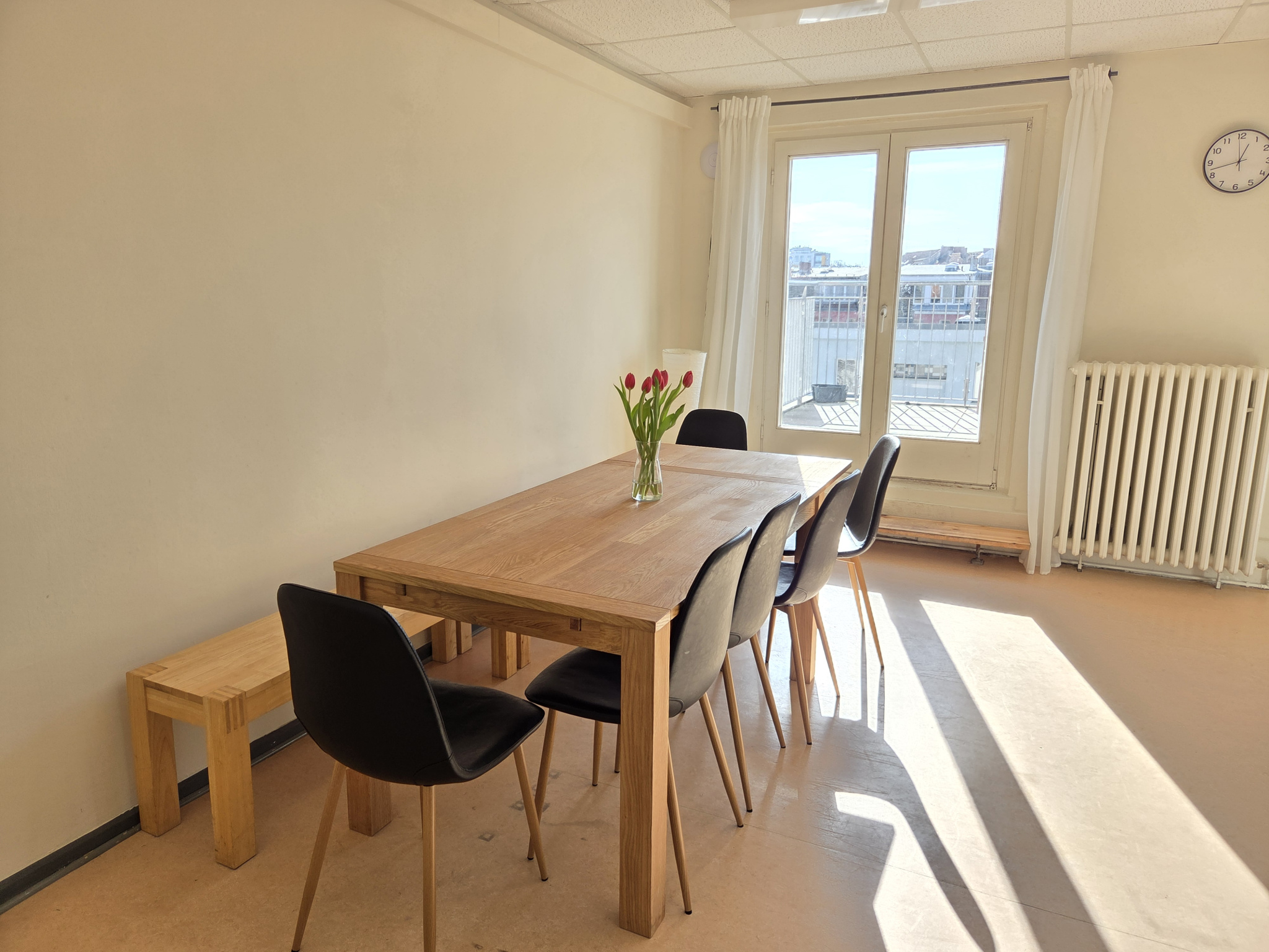 Ein Holztisch mit einer Sitzbank und fünf Stühlen in einem hellen Zimmer mit einer Terassentür nach draußen. Auf dem Tisch steht eine Vase mit frischen Blumen