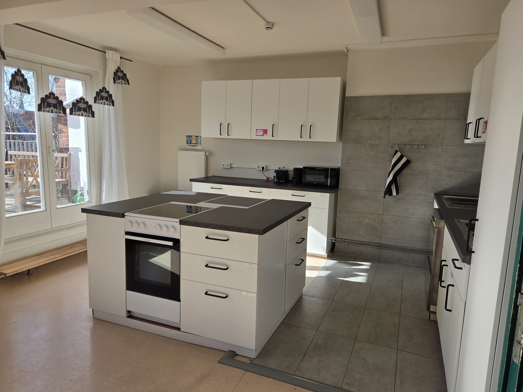 Eine helle Küche mit weißen Schränken und grauen Arbeitsflächen und einer Terassentür nach draußen