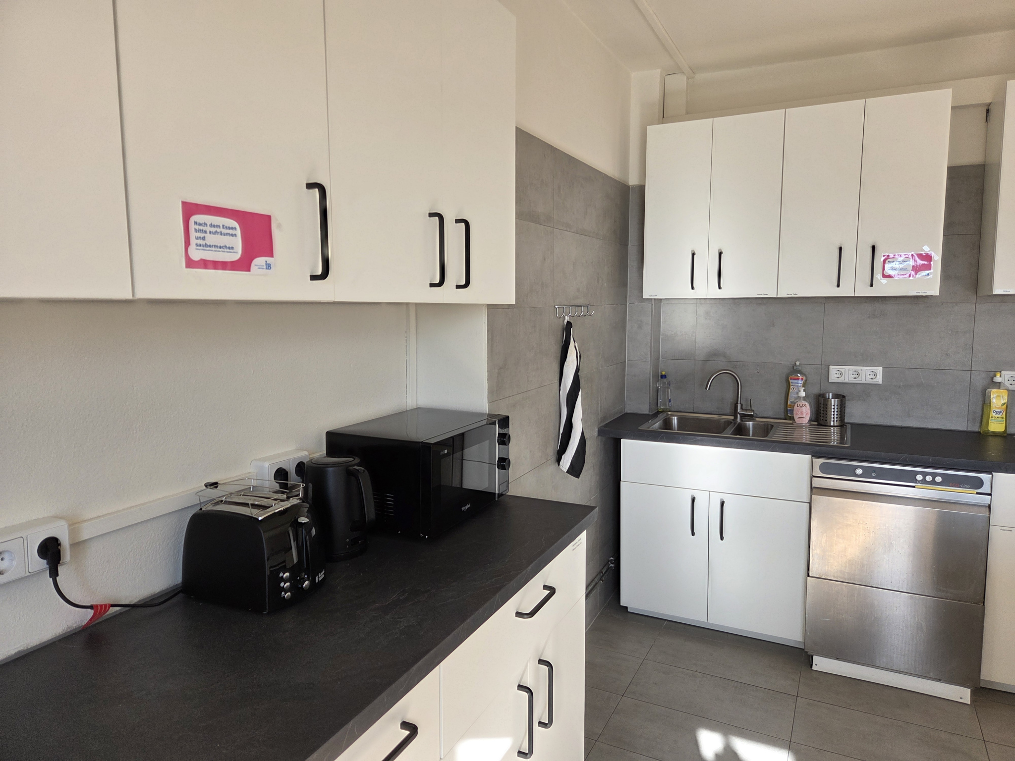 Eine helle Küche mit weißen Schränken und grauen Arbeitsflächen, ein Toaster, eine Mikrowelle, eine Spülmaschine.