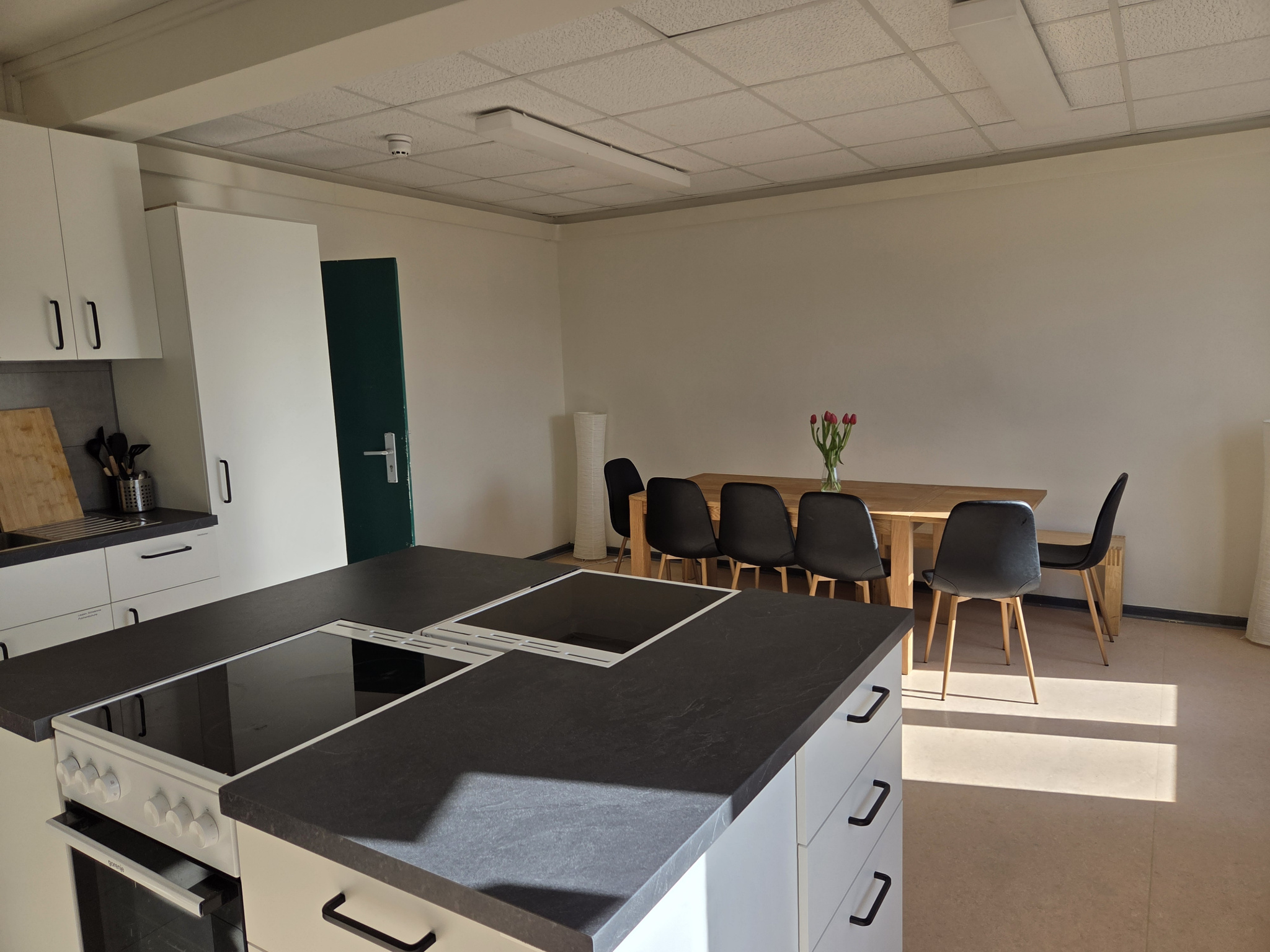 Eine helle Küche mit weißen Schränken und grauen Arbeitsflächen und einem Holztisch mit einer Sitzbank und fünf Stühlen. Darauf steht eine Blumenvase mit frischen Blumen.