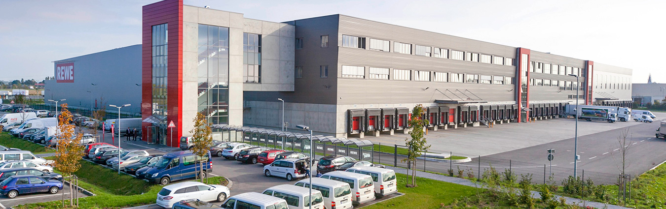 Großes REWE-Logistikzentrum mit moderner Architektur, mehreren Laderampen, Lieferfahrzeugen und Parkplatz im Vordergrund