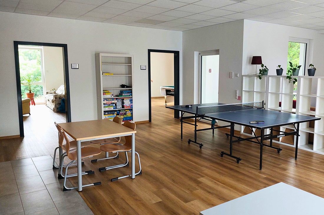 Ein großer, heller Raum mit einer Tischtennisplatte und einem weißen Kalax Regal, einem Tisch mit vier Stühlen und einem weiteren Regal mit Spielen.