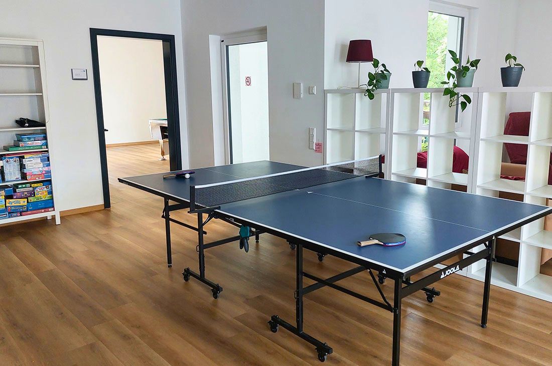 Ein großer, heller Raum mit einer Tischtennisplatte und einem weißen Kalax Regal und einem weiteren Regal mit Spielen. Es führen zwei Türen aus dem Raum hinaus. 