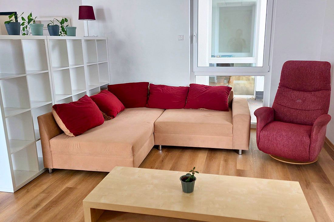 Eine weißes Kalaxregal neben einem beigen Sofa mit roten Kissen, einem roten Sessel und einem Couchtisch.