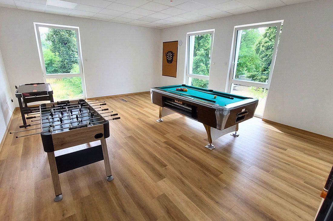 Ein großer heller Raum mit Billardtisch, ein Kickertisch, ein Air Hockey Tisch und einer Dart Scheibe an der Wand.