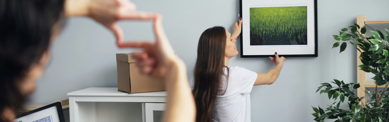Eine Person hängt ein gerahmtes Bild mit einer grünen Landschaft an eine Wand. Im Vordergrund zeigt eine unscharfe Hand die Position des Bildes an. Im Raum stehen Umzugskartons, ein Regal mit Pflanzen und weitere Bilderrahmen. Die Szene vermittelt das Einrichten einer neuen Wohnung.