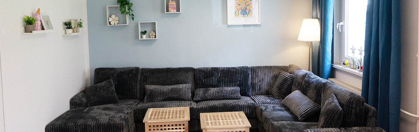Gemütlicher Wohn- und Aufenthaltsraum mit großer dunkler Eckcouch, zwei Beistelltischen, Stehlampe, dekorativen Wandregalen und Fenster mit blauen Vorhängen.