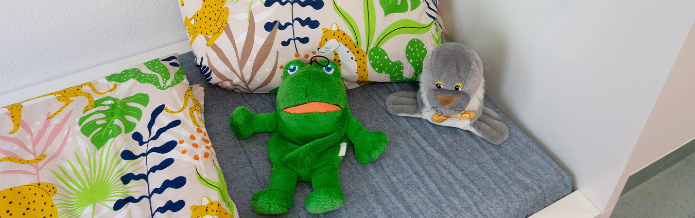 Kuschelige Sitzecke mit bunten Kissen im Dschungel-Design sowie einem grünen Frosch-Kuscheltier und einem grauen Vogel-Kuscheltier auf einer Sitzbank.